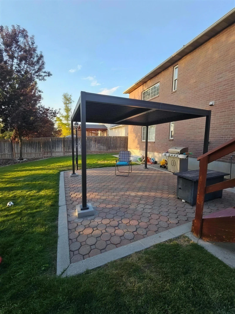 Finished black louvered aluminum pergola over a brick paver patio beside a two-story brick home