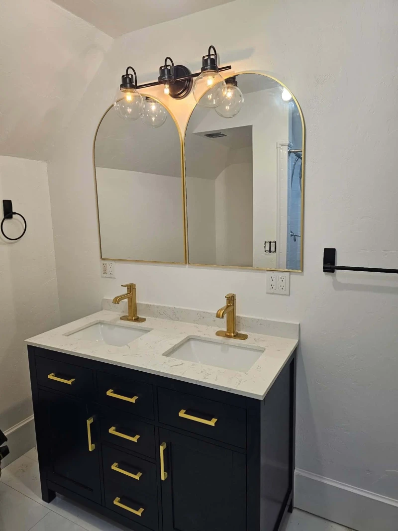 Finished bathroom vanity with twin arched gold-framed mirrors, a three-globe black vanity light, and brushed gold faucets on the marble-look top